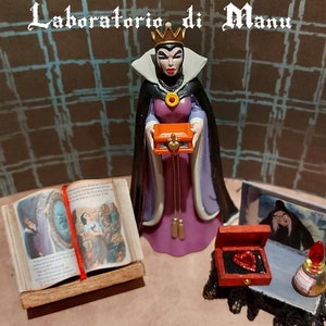 Puede incluir: Una figura de la Reina Malvada de Blancanieves sosteniendo una pequeña caja naranja con un corazón en ella. Hay un libro con una imagen de espejo a la izquierda, una pequeña caja roja con un corazón rojo a la derecha y una botella de poción roja.