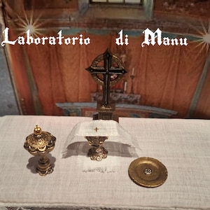 May include: A miniature altar set with a gold chalice, paten, and ciborium on a white linen tablecloth with a lace trim. The altar is decorated with a black metal cross.