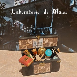 Puede incluir: Dos cofres del tesoro de madera negra, uno abierto y lleno de botellas de pociones en miniatura, un libro rojo, un colgante de sol dorado y una bola de cristal azul. El texto "Laboratorio di Manu" está encima de los cofres.