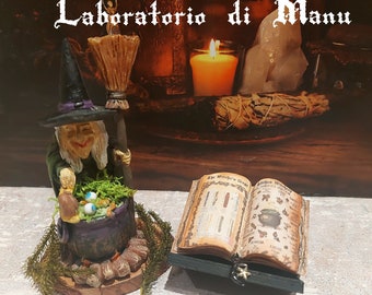 Puede incluir: Una figura caprichosa de bruja con sombrero negro y túnica verde, revolviendo un caldero. Junto a ella, un libro abierto sobre un soporte. La imagen incluye una vela encendida y el texto "Laboratorio di Manu".