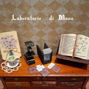 Puede incluir: Un conjunto de laboratorio en miniatura con un microscopio, un libro sobre Matricaria Chamomilla y una pequeña taza de té sobre una mesa de madera con cajones. El libro contiene una ilustración botánica de la planta y un texto sobre sus propiedades medicinales. El texto del libro dice: "Esta especie de manzanilla se utiliza para fines medicinales y cosméticos. El extracto de manzanilla se utiliza en productos cosméticos, especialmente en cremas y la infusión de enjuague para el cabello, les da un brillo dorado. La manzanilla de farmacia también es conocida por sus propiedades calmantes, sedantes, antiespasmódicas, antisépticas y antieméticas. Matricaria Chamomilla"