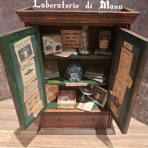 Può includere: Un mobiletto in legno in miniatura con ante aperte etichettato Laboratorio di Manu, contenente piccoli oggetti di scena tra cui un cartello Project Orion, una figura in una teca di vetro, una sfera geometrica, libri e vari documenti con testo come Top Secret e Unidentified Aerial Phenomena.