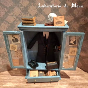 Può includere: Un armadietto in miniatura in legno azzurro con ante aperte, contenente una giacca da completo nera, un paio di scarpe nere, una piccola valigetta marrone e libri. Il mobile presenta un cassetto, un teschio bianco e varie stampe su carta.