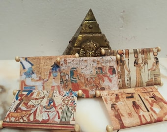 May include: Five miniature scrolls with Egyptian hieroglyphics and images of gods and goddesses. The scrolls are displayed on a white surface with a small gold pyramid in the background.