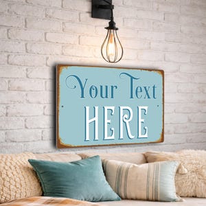 May include: A rectangular metal sign with a light blue background and a distressed brown border. The sign features the text "Your Text HERE" in a decorative white and dark blue font. The sign has small holes in the corners for mounting.