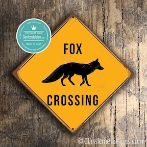 May include: A yellow diamond-shaped metal sign with a black silhouette of a fox and the words "FOX CROSSING" in black. The sign has a distressed, vintage look and is mounted on a wooden surface. The sign also has a round logo in the upper left corner.