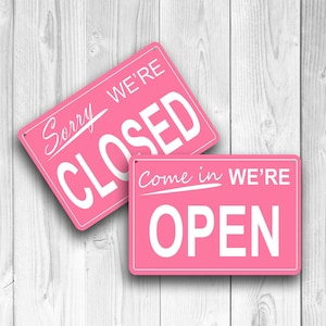 Peut inclure: Deux panneaux rectangulaires roses avec du texte blanc. L'un des panneaux indique "Sorry We're Closed" et l'autre "Come in We're Open". Les panneaux sont placés sur un fond en bois blanc.