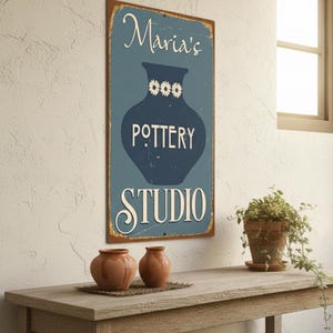 Può includere: Un'insegna rettangolare con la scritta "Maria's Pottery Studio" in bianco. Lo sfondo è blu con un vaso blu scuro. Due piccoli vasi di ceramica marrone sono su un tavolo di legno.