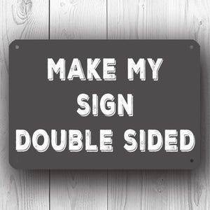 May include: A dark grey metal sign with the white text "MAKE MY SIGN DOUBLE SIDED". The sign is rectangular with rounded corners and is mounted on a light grey wooden background. The text is in bold, uppercase letters.