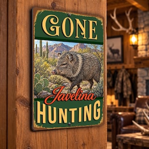 May include: A rectangular metal sign with a dark green border and a distressed finish. The sign features the text GONE at the top and JAVELINA HUNTING at the bottom in yellow and orange. A central illustration depicts a javelina in a desert.