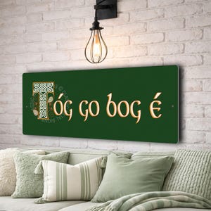 Könnte beinhalten: Rechteckiges, dunkelgrünes Metallschild mit goldfarbenen Buchstaben und keltischem Design. Das Schild lautet "Tóg go bog é" und ist an einer Ziegelmauer über einem Sofa mit grünen Kissen befestigt.