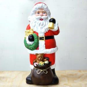 May include: A large Santa Claus figurine, wearing a red suit with white trim, a black belt, and boots. He holds a gold bell and a green wreath. A brown sack at his feet is filled with gifts and a teddy bear. Christmas decor.