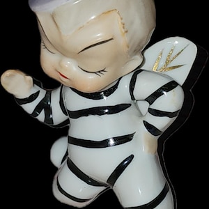May include: A vintage ceramic figurine of a cherubic figure in a sailor suit with black and white stripes. The figurine has closed eyes, a small red mouth, and a white hat. The wings are white with gold accents.