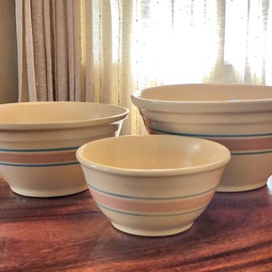 May include: Three off-white ceramic mixing bowls of varying sizes. Each bowl has a horizontal stripe design in alternating colors of light blue and pink. The bowls are arranged on a wooden surface.