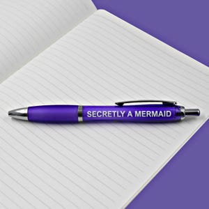 May include: A purple pen with the text "Secretly a Mermaid" printed on it. The pen is lying on a lined notebook.