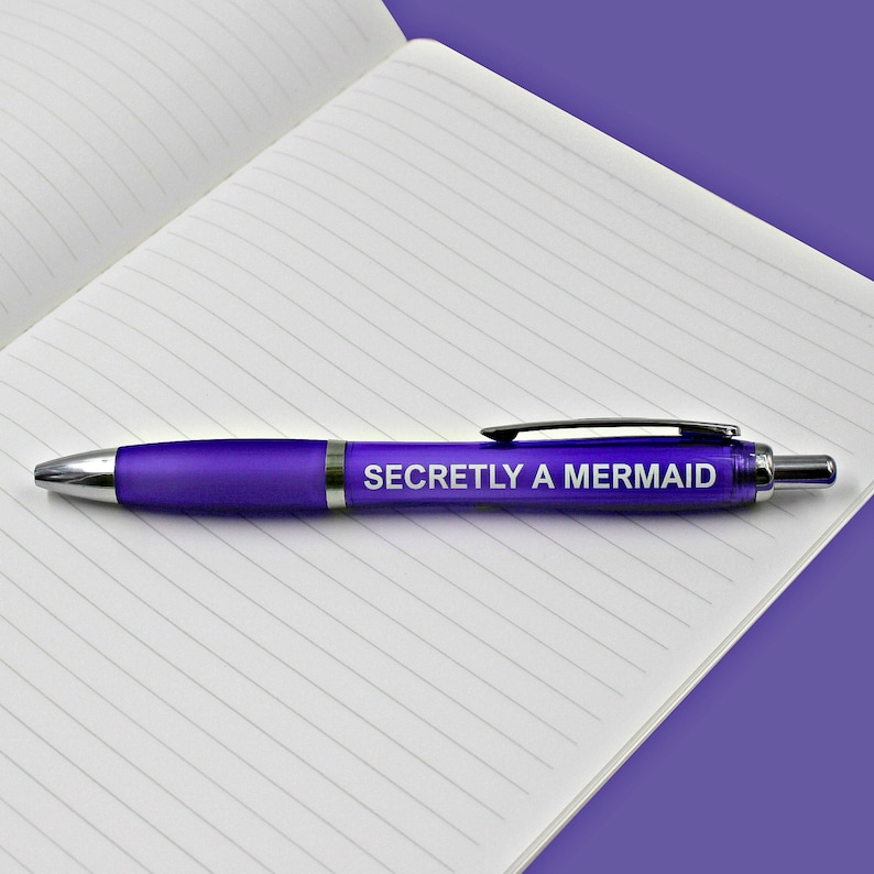 May include: A purple pen with the text "Secretly a Mermaid" printed on it. The pen is lying on a lined notebook.