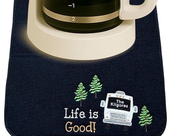 May include: A dark blue rectangular fabric mat featuring embroidered green pine trees, a white recreational vehicle with the text The Kilgores, and the embroidered phrase Life is Good in white and tan, positioned under a coffee carafe base.