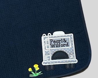 May include: Dark navy blanket with a textured surface. It features an embroidered design of a grey camper with the text "Pearl & Wilford" and a small yellow flower. The camper has white polka dots.