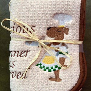May include: A beige kitchen towel with brown trim and a brown embroidered dog wearing a chef's hat. The towel has the text "Serious Dinner is Served" embroidered on it.