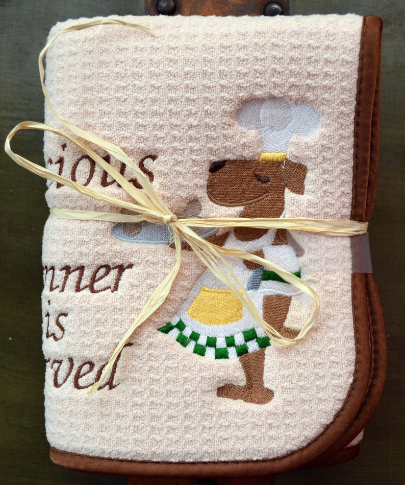 May include: A beige kitchen towel with brown trim and a brown embroidered dog wearing a chef's hat. The towel has the text "Serious Dinner is Served" embroidered on it.