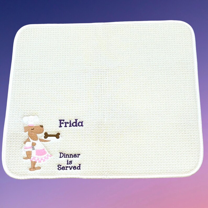 May include: White placemat with a brown dog wearing a chef's hat and a pink apron. The dog is holding a bone and the text "Frida Dinner is Served" is embroidered on the placemat.
