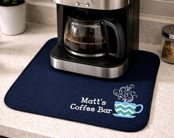 May include: A navy blue coffee bar mat with the text "Matt's Coffee Bar" and a coffee cup design. A coffee machine sits on the mat. The coffee cup design has a blue and green chevron pattern.