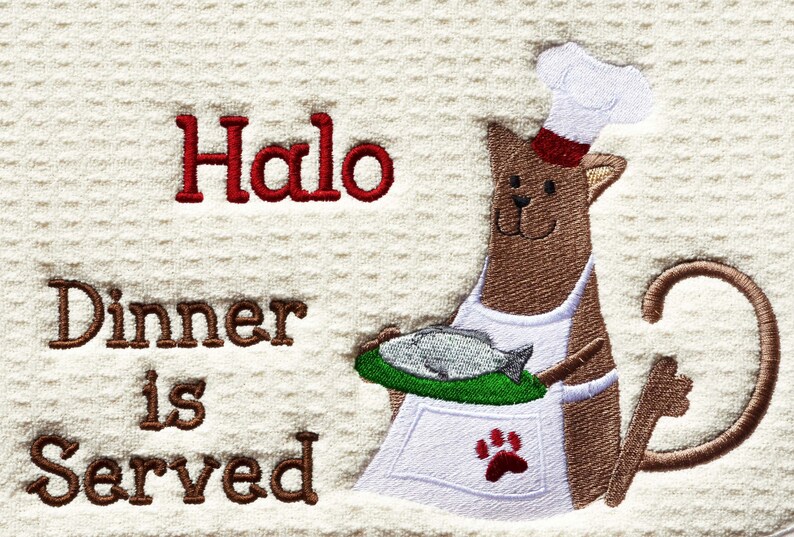 May include: A brown cat wearing a white chef's hat and apron holds a plate with a fish on it. The text "Halo Dinner is Served" is embroidered on the white background.