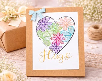 May include: A handmade greeting card featuring a heart-shaped floral design in purple, pink, green, and orange. The word "Hugs" is written in gold script below the heart. A small blue bow is attached to the top of the card.