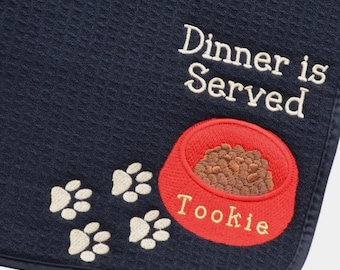 May include: A dark navy blue waffle-textured mat featuring the embroidered white text "Dinner is Served" above a red pet bowl filled with brown kibble. The bowl is labelled "Tookie" in white, accompanied by four white paw prints.