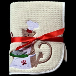 May include: A cream-colored kitchen towel with a red ribbon tied around it. The towel features an embroidered design of a brown cat wearing a chef's hat and holding a pan of fish.