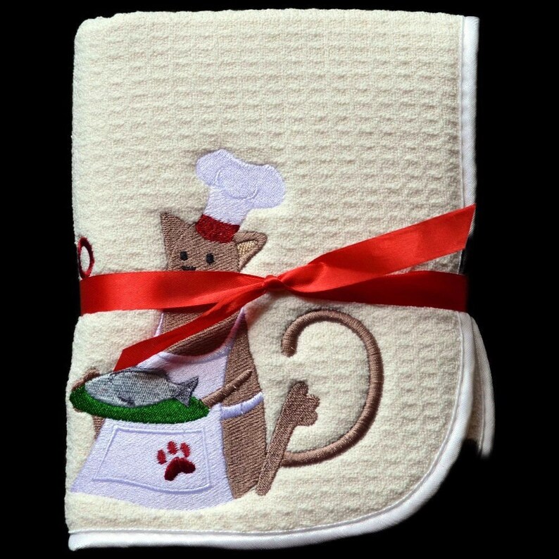 May include: A cream-colored kitchen towel with a red ribbon tied around it. The towel features an embroidered design of a brown cat wearing a chef's hat and holding a pan of fish.