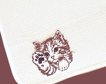May include: White, textured towel featuring a brown embroidered illustration of a kitten holding its paw up. The kitten has brown and white striped fur and large eyes. The towel has a rounded corner.