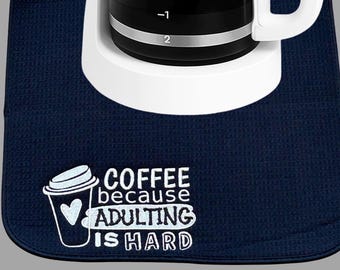 May include: A navy blue coffee machine mat with the text "COFFEE because ADULTING IS HARD" in white. A white coffee maker sits on the mat. The mat has a textured, waffle-like pattern.