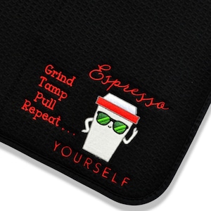 May include: Black kitchen towel with embroidered text and a coffee cup graphic. The red text reads "Grind, Tamp, Pull, Repeat... Espresso YOURSELF." The coffee cup has sunglasses and a red lid.