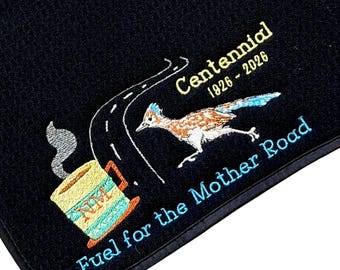 May include: A black fabric mat featuring an embroidered design of a roadrunner, a steaming coffee mug with NM text, and a winding road. The text reads Centennial 1926-2026 and Fuel for the Mother Road in yellow and light blue thread colours.