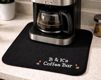 May include: A black coffee bar mat with the text "B & K's Coffee Bar" in white, along with two coffee bean illustrations. A silver and black coffee maker with a glass carafe sits on the mat. The time displayed on the coffee maker is 8:00.