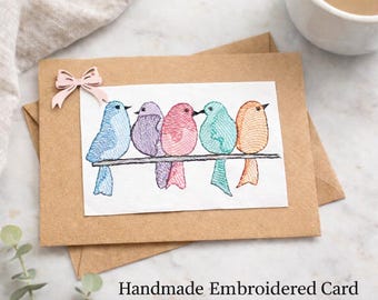 May include: A rectangular kraft paper greeting card featuring an embroidered white fabric patch with five colourful birds perched on a line. The birds are blue, purple, pink, teal, and orange. A small light pink paper bow is attached to the top left.