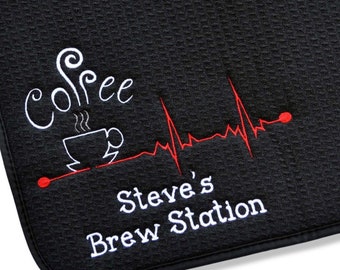 Puede incluir: Una alfombrilla de secado negra texturizada con un icono de taza de café bordado en blanco y la palabra Coffee encima. Una línea roja de ritmo cardíaco EKG cruza la alfombrilla. Debajo, el texto Steve's Brew Station está bordado en blanco.
