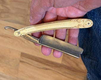 May include: A vintage straight razor with a gold-colored handle and blade. The handle features ornate floral designs and a textured edge. The blade has engraved text. A classic grooming tool.