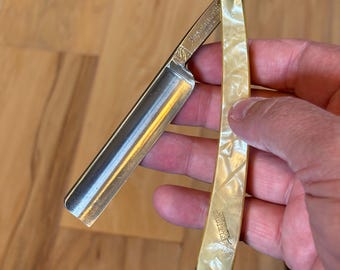 May include: A straight razor with a silver blade and a cream-colored handle. The handle has a pearlescent finish and is marked with the word "PLEIADES". The blade is open and ready for use.
