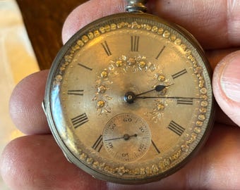 May include: Antique pocket watch with a silver-tone case and a cream-colored face. The watch features Roman numeral hour markers, delicate floral detailing, and a smaller subdial for seconds. The watch has a loop at the top for attaching a chain.