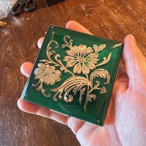 May include: A square, emerald green compact with a silver floral design. The compact is held in a hand and has a shiny, reflective surface. The floral design features two large flowers and intricate leaf patterns.