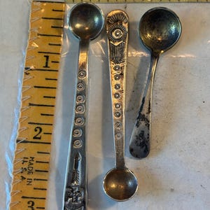 May include: Three silver-toned metal spoons of varying designs placed next to a yellow measuring tape. The left spoon features circular patterns and an arrow motif. The center spoon has a geometric top and circular details. The right spoon is plain.