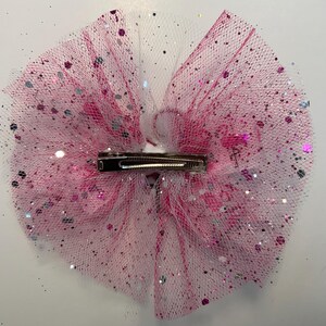 May include: A pink and white tulle hair bow with silver and pink glitter accents. The bow features a metal clip on the back for easy attachment. This accessory is perfect for adding a touch of sparkle to any hairstyle.
