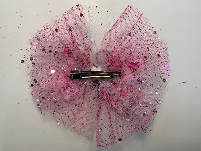 May include: A pink and white tulle hair bow with silver and pink glitter accents. The bow features a metal clip on the back for easy attachment. This accessory is perfect for adding a touch of sparkle to any hairstyle.