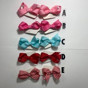 May include: A collection of hair bows in various colors, including pink, light blue, red, and a red and white gingham pattern. Each bow is attached to a small card and labeled with a letter from A to E.