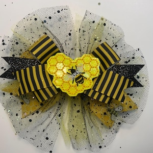 May include: A decorative hair bow featuring a bee theme. The bow has black and yellow striped ribbons, black glitter accents, and a yellow honeycomb-shaped center with a bee applique. The bow is set on a black mesh base with black speckles.