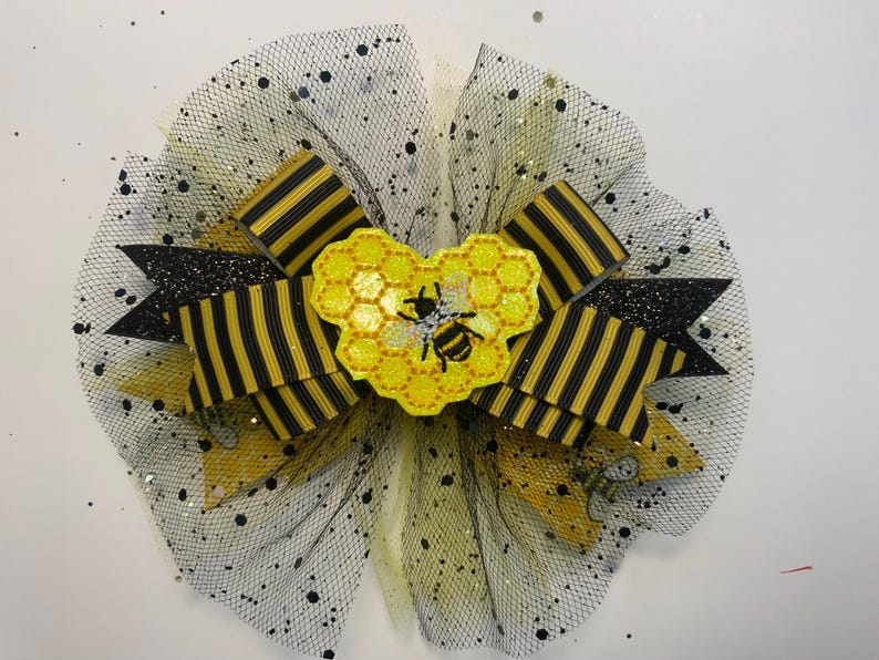 May include: A decorative hair bow featuring a bee theme. The bow has black and yellow striped ribbons, black glitter accents, and a yellow honeycomb-shaped center with a bee applique. The bow is set on a black mesh base with black speckles.