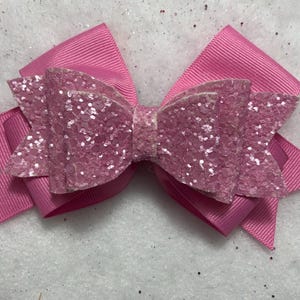 May include: A pink glitter bow with a large center bow and two smaller side bows. The bow is made of pink ribbon and has a sparkly finish.