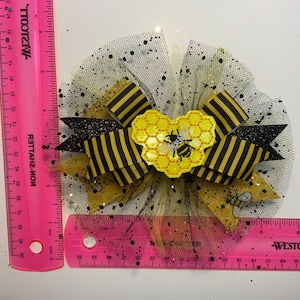 May include: A decorative hair bow featuring a bee theme. The bow has black and yellow striped ribbon, a yellow honeycomb heart with a bee, and a black tulle base with glitter accents. The bow measures approximately 4 inches in diameter.